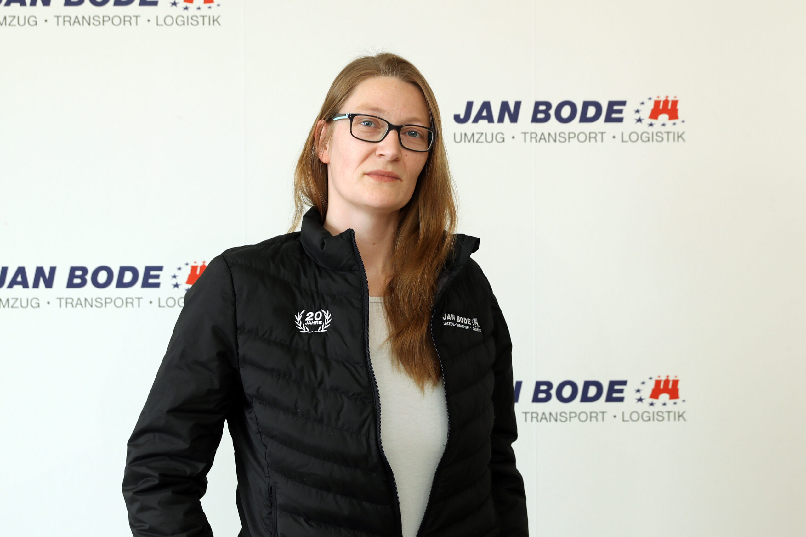Bild von der Mitarbeiterin Alexandra Vauk vor einer weißen Wand mit dem Logo von der Spedition Jan Bode.